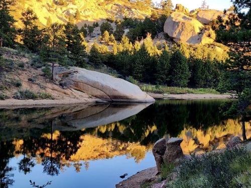 A serene landscape featuring a calm lake reflecting golden rocks and lush green trees under a clear sky.