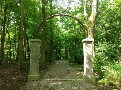 A gravel path through a lush green forest, marked by stone pillars and archways overhead.