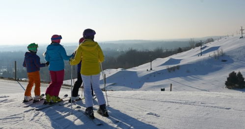 Four skiers in colorful outfits stand on a snowy slope, overlooking a winter landscape under a clear sky.