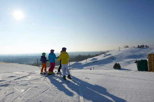 Four children in colorful ski gear stand on a snowy slope, looking out at a clear blue sky and distant hills.
