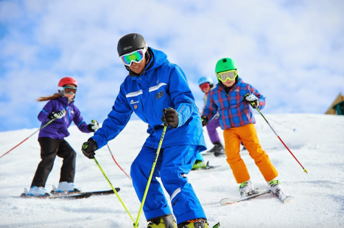 A ski instructor guides four children on a snowy slope, all wearing colorful ski gear and helmets under a blue sky.