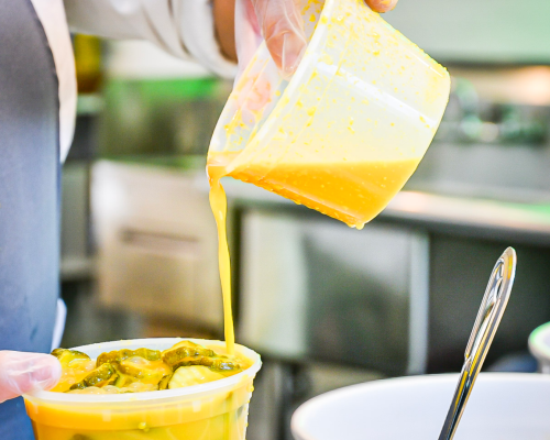 A chef pours a creamy yellow sauce over a container of pickles in a kitchen setting.