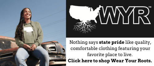 A smiling woman in a hoodie sits on a vintage truck, promoting state pride clothing with the slogan "Wear Your Roots."