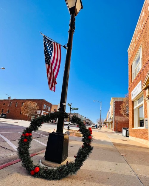 A street scene featuring a lamppost with an American flag and a festive wreath on the ground, under a clear blue sky.