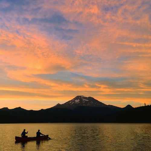 Silhouetted figures in a canoe on a lake at sunset, with mountains and colorful clouds in the background.
