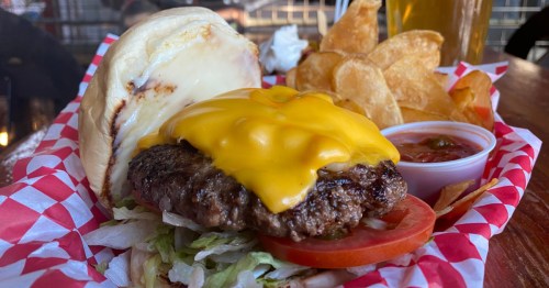 The top bun of a buffalo burger sits to the side of a patty covered in cheese. A handful of chips sits next to the burger.