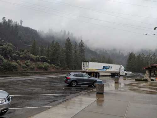 A truck parked in a misty, rainy lot surrounded by trees and mountains. Wet pavement reflects the cloudy sky.