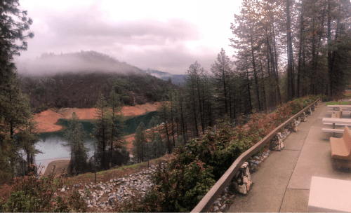 A serene landscape featuring a lake surrounded by trees and misty mountains, with a pathway and seating area in the foreground.