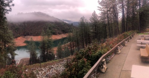 A scenic view of a misty lake surrounded by trees, with a pathway and seating area in the foreground.