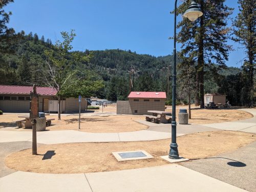 A sunny park scene with picnic tables, a path, and trees, set against a backdrop of green hills.