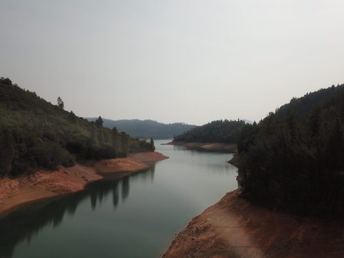 A serene lake surrounded by forested hills, with calm waters reflecting the landscape under a hazy sky.