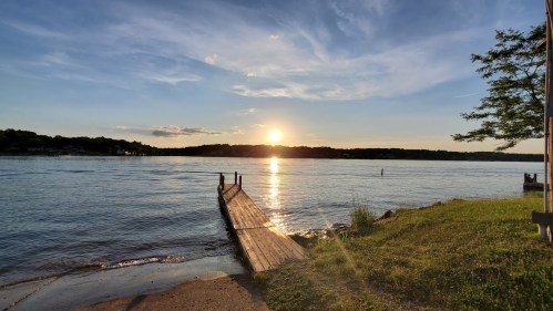 A serene lakeside view at sunset, with a wooden dock extending into calm waters and a grassy shore.