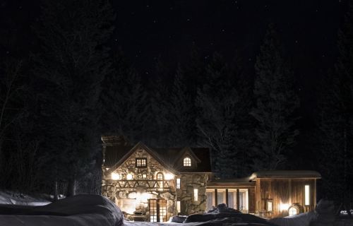 A cozy stone cabin illuminated at night, surrounded by snow and tall trees under a starry sky.