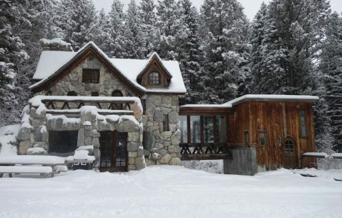 A snow-covered stone and wood cabin surrounded by tall evergreen trees in a winter landscape.