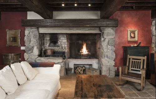 Cozy living room with a stone fireplace, wooden beams, and rustic furniture, featuring a warm fire and red walls.