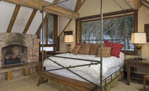 Cozy bedroom with a four-poster bed, stone fireplace, and large windows overlooking a snowy landscape.