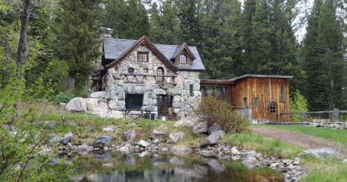 A charming stone cottage surrounded by trees, with a wooden addition and a small pond in the foreground.