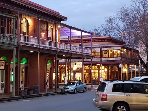 Historic buildings line a street, featuring shops and restaurants, with evening lights illuminating the scene.
