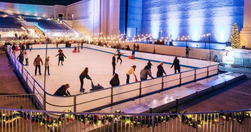 A festive ice skating rink with people skating, surrounded by lights and holiday decorations.