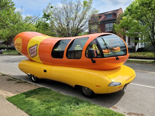 A colorful Oscar Mayer Wienermobile parked on a street, surrounded by trees and residential buildings.