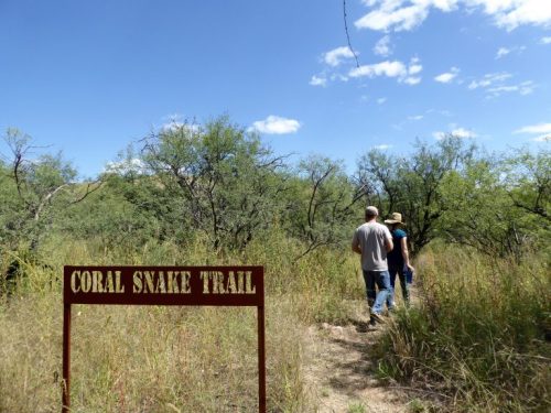 Two people walk along a grassy path marked "Coral Snake Trail" surrounded by trees and blue skies.