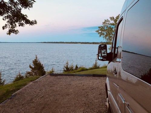 A serene lakeside view with a van parked on a gravel area, surrounded by trees and a calm water surface.