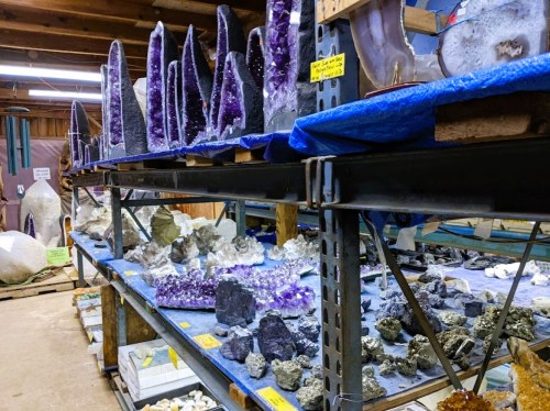 A display of various crystals and geodes on shelves in a well-lit warehouse, showcasing different colors and shapes.