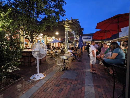 A lively outdoor dining area at dusk, featuring tables, umbrellas, and string lights, with people enjoying the atmosphere.