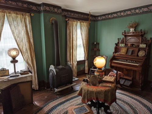 A vintage room with green walls, an antique piano, a wood stove, and decorative curtains, featuring a round rug and lamps.