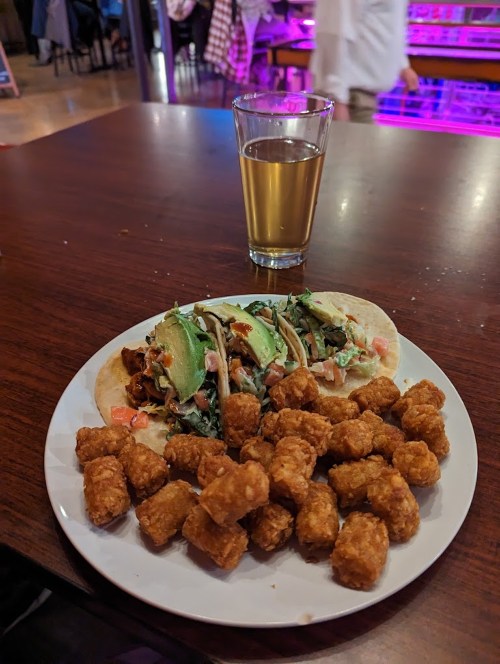 A plate with two tacos topped with avocado and greens, alongside a serving of crispy tater tots and a glass of drink.