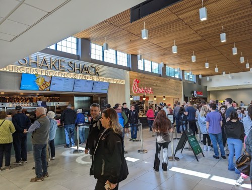 A busy food court with long lines at Shake Shack and Chick-fil-A, featuring a modern interior and large windows.