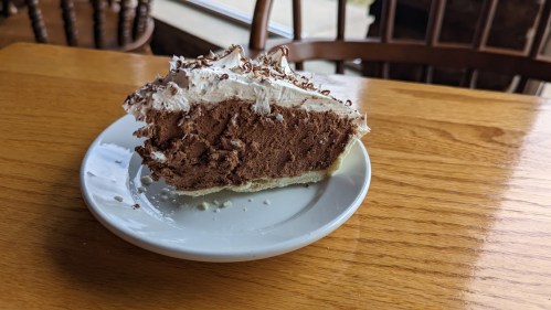 A slice of chocolate cream pie with whipped cream on a white plate, resting on a wooden table.