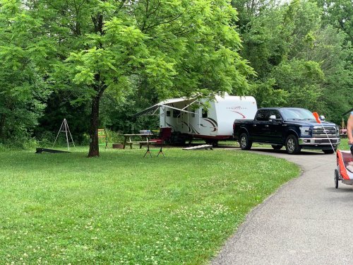 A black truck parked next to a travel trailer under a green tree, with a picnic table nearby in a grassy area.