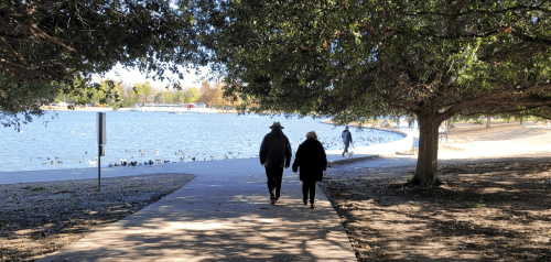 Two people walk along a path by a lake, surrounded by trees, with ducks visible in the water.