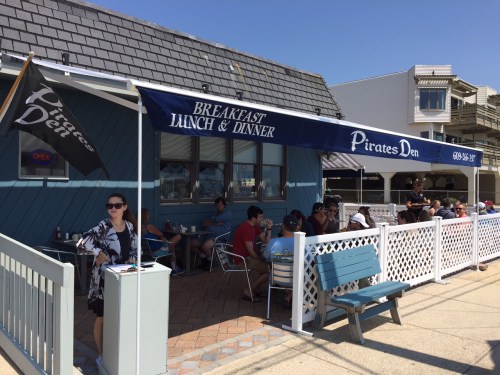 A seaside restaurant, Pirates Den, with outdoor seating and a flag, bustling with diners enjoying breakfast and lunch.