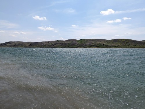 A serene view of a calm sea with gentle waves, bordered by green hills under a clear blue sky.