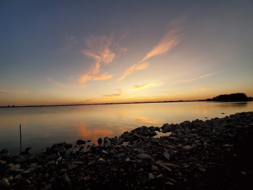 A serene sunset over a calm lake, with colorful clouds reflecting on the water and rocky shoreline in the foreground.