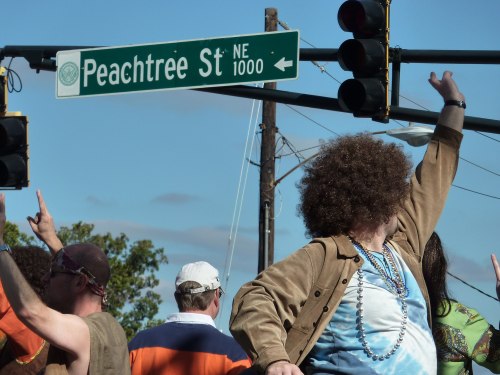 A crowd celebrates at a street corner with a Peachtree St NE sign, featuring a person with a large curly hairstyle.