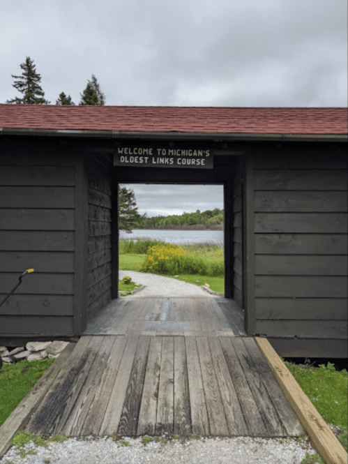 A wooden entrance with a sign reading "Welcome to Michigan's Oldest Links Course," leading to a scenic view.