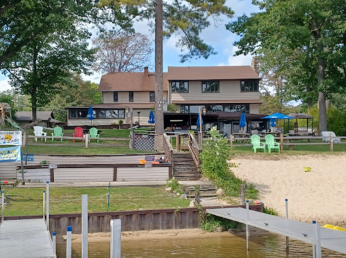 A lakeside view of a house with a sandy beach, colorful chairs, and a dock under a clear blue sky.