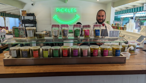 A smiling vendor stands behind a counter filled with jars of pickles and condiments, with a neon "Pickles" sign above.
