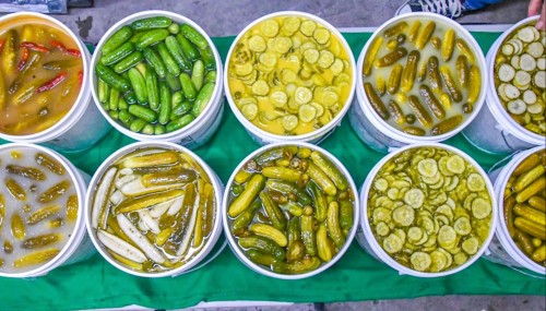 Aerial view of various buckets filled with pickled vegetables, including cucumbers and peppers, arranged in two rows.