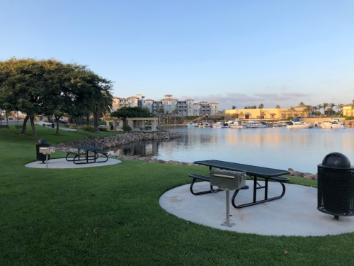 A serene park by the water featuring picnic tables, a grill, and boats in the background under a clear sky.