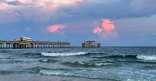 A serene beach scene with gentle waves, a pier, and colorful clouds at sunset.