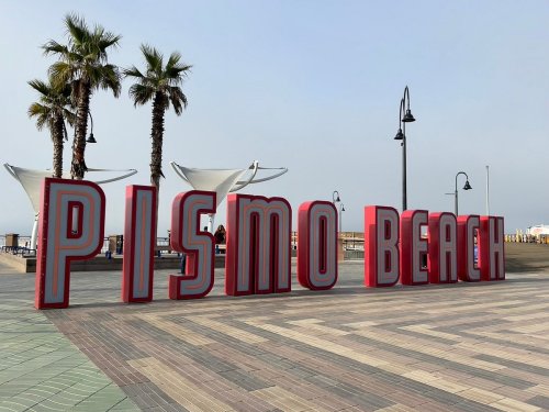 Colorful "PISMO BEACH" sign on a boardwalk, with palm trees and a cloudy sky in the background.