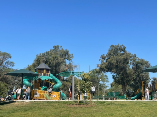A sunny playground with green slides and structures, surrounded by trees and people enjoying the space.