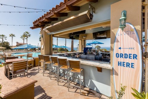 A poolside bar with a surfboard sign reading "Order Here," surrounded by palm trees and blue umbrellas.