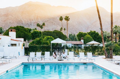 A serene poolside scene with two people relaxing near a sparkling pool, surrounded by palm trees and mountains at sunset.