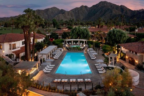 Aerial view of a resort pool surrounded by palm trees and mountains at sunset, with lounge chairs arranged around the pool.