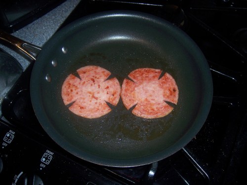 Two slices of sausage cooking in a frying pan, with decorative cuts creating a flower-like shape.